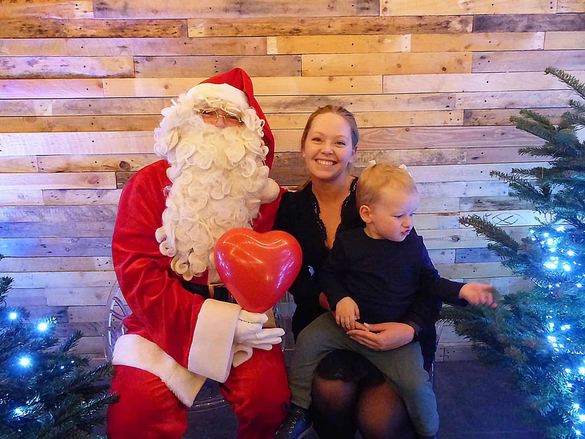 Père Noël portant un ballon en forme de cœur, assis à côté d’une jeune femme tenant son petit garçon sur ses genoux.